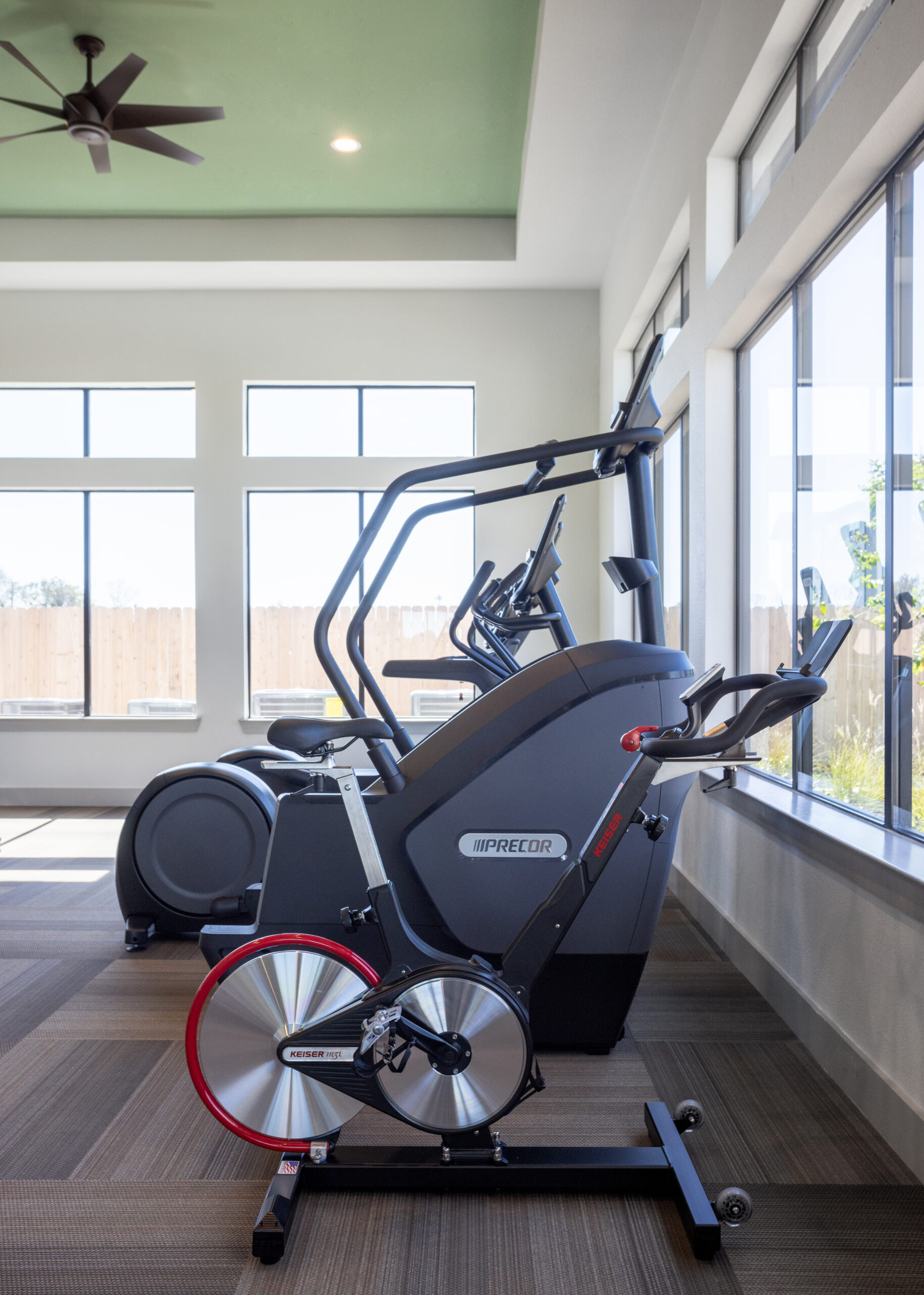 indoor fitness center with free weights, treadmills, bicycle/bike, with large window in the back shining bright light in at prose evergreen apartments in new braunfels, texas